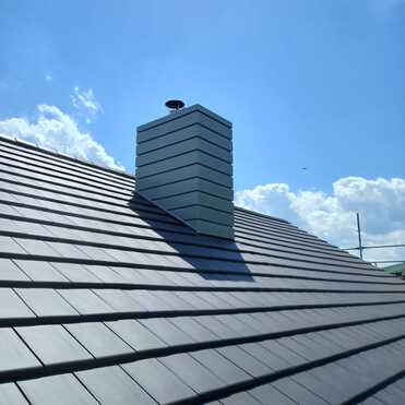 Moderner Schornstein mit Metallverkleidung auf frisch gedecktem Ziegeldach unter blauem Himmel.