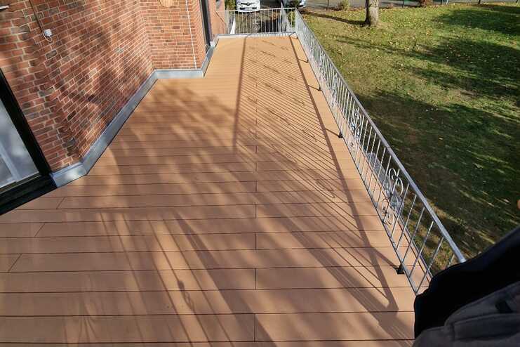 Langer, rechteckiger Balkon eines Wohngebäudes mit hellbraunen Bodenfliesen und einem schmiedeeisernen Geländer, mit Blick auf einen baumbestandenen Parkplatz an einem sonnigen Herbsttag.