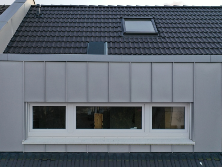 Ansicht einer modernen Dachgaube mit großen Fenstern und grauen Paneelen, eingebettet in ein dunkles Schieferdach eines Wohnhauses.