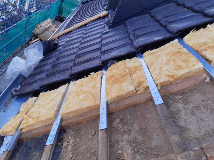Detailaufnahme eines Dachteils während der Renovierung, zeigt neue gelbe Isoliermatten zwischen Holzlatten und teilweise verlegte dunkle Dachziegel.
