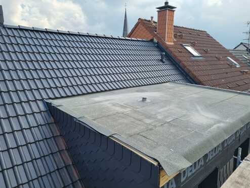 Überblick über ein graues Flachdach neben einem geneigten Dach mit dunkelgrauen Schindeln und einem roten Ziegeldach mit Dachfenstern im Hintergrund.