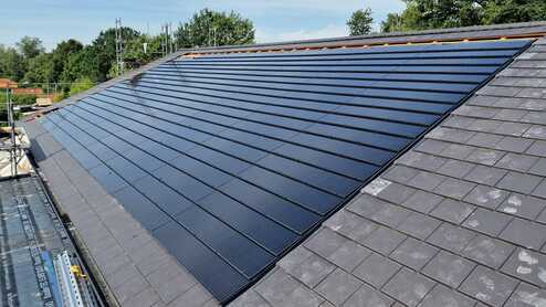 Große Fläche von Solarpaneelen auf einem Schrägdach neben dunkelgrauen Schieferziegeln, unter blauem Himmel mit Bäumen im Hintergrund.