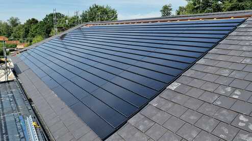 Große Fläche von Solarpaneelen auf einem Schrägdach neben dunkelgrauen Schieferziegeln, unter blauem Himmel mit Bäumen im Hintergrund.