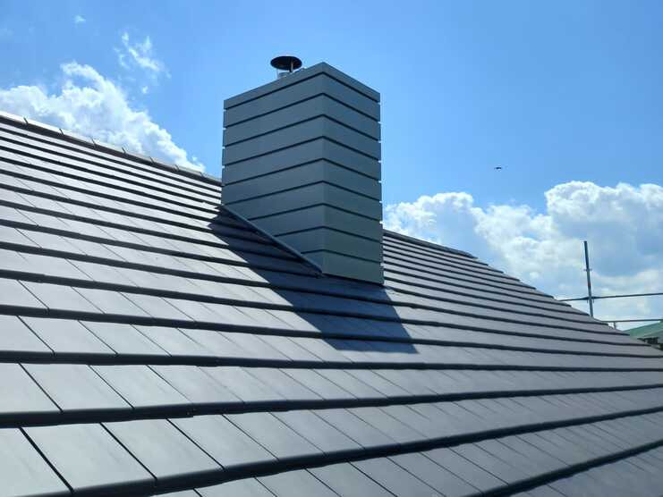 Ein modern gestalteter Schornstein aus gestreiften Metallpaneelen ragt aus einem dunkelgrauen Ziegeldach heraus. Klare blaue Himmel mit wenigen Wolken im Hintergrund.